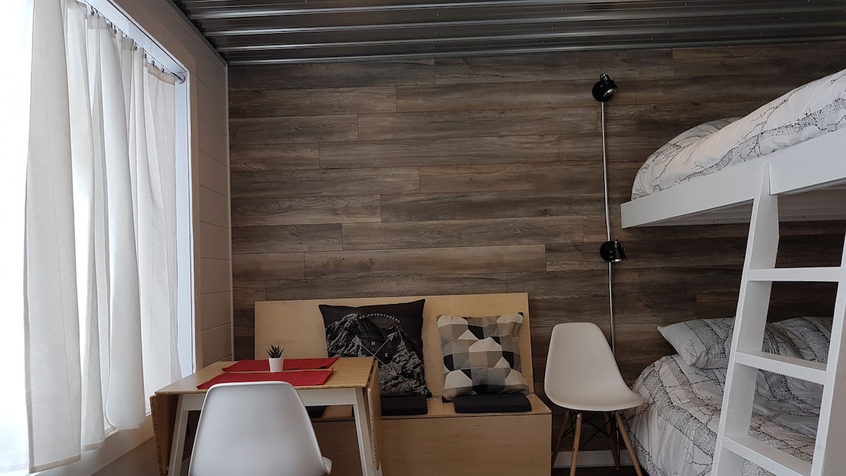 A cozy interior features a bunk bed with neatly arranged bedding. A small wooden table is set with a red placemat, while two white chairs accompany it. A light fixture and decorative cushions enhance the space, and natural light filters through a sheer curtain.