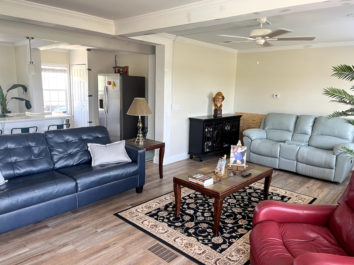 A welcoming living space features a mix of seating options, including a stylish blue couch and a vibrant red armchair. A wooden coffee table sits atop a patterned area rug, complemented by a decorative lamp. The open layout connects seamlessly to the dining area in the background.
