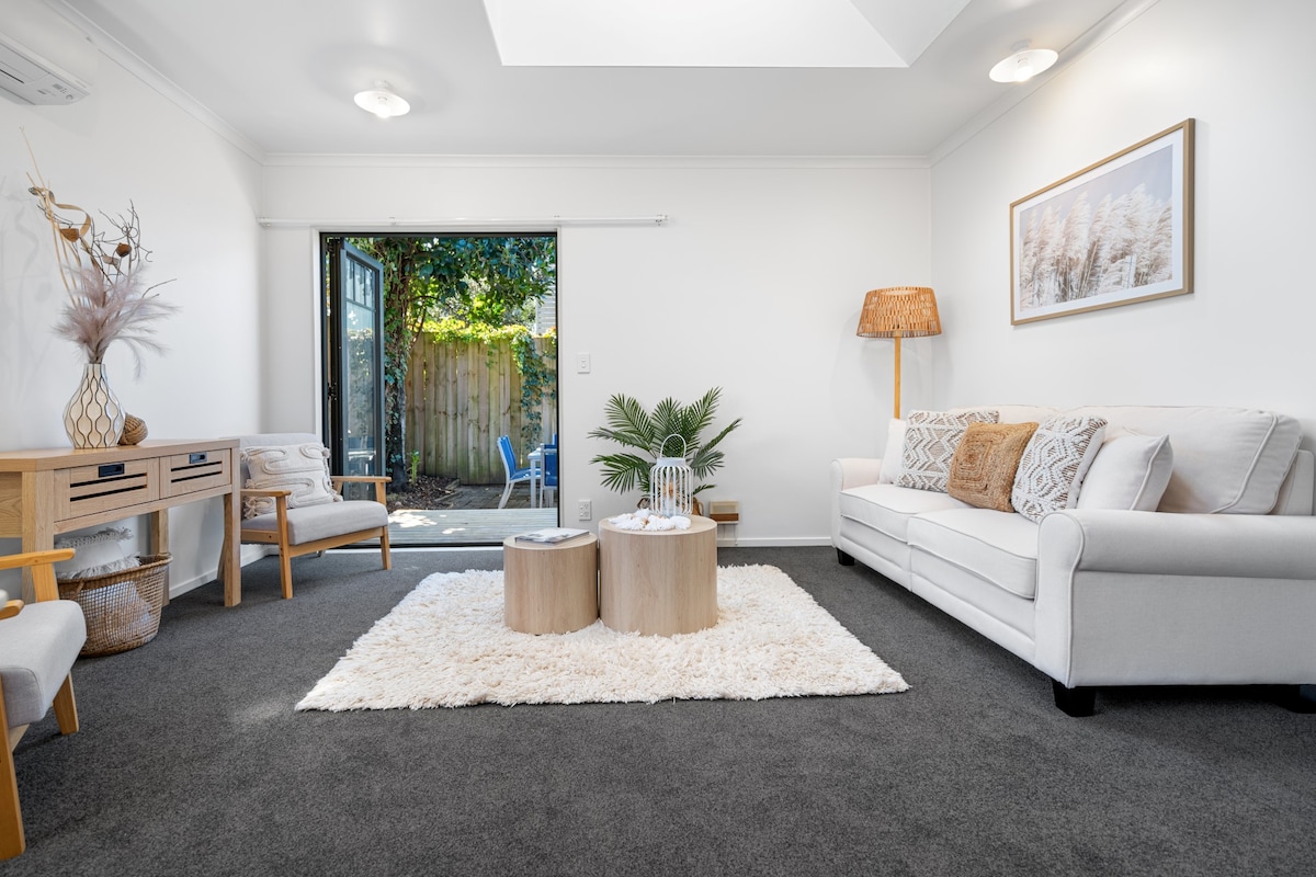 A cozy living area is presented with light-colored furniture, including a sofa and accent chairs. A soft rug covers the dark carpet, while two wooden tables sit in the center. Natural light enters through a sliding glass door that leads to a garden.