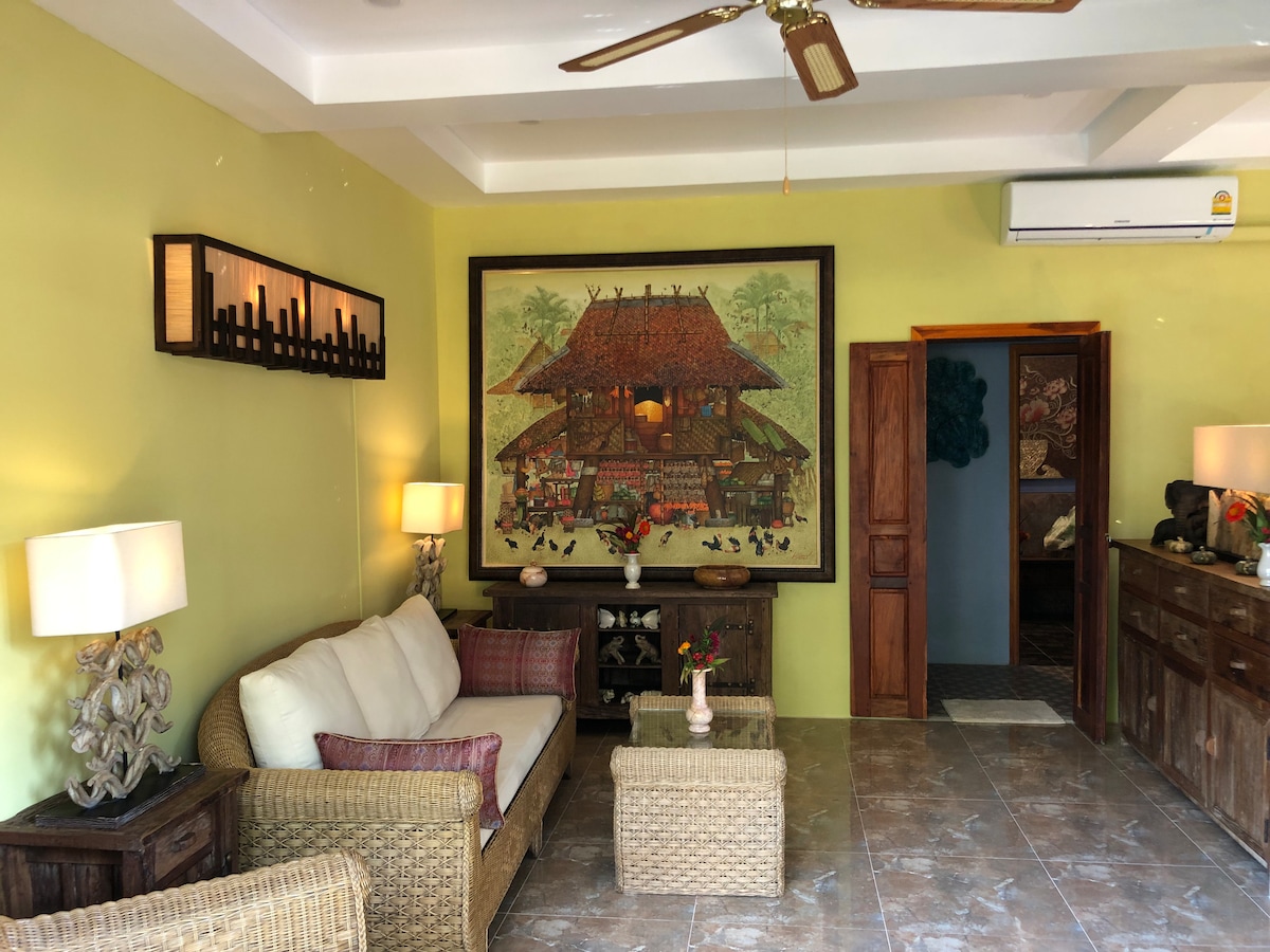 A cozy living area features a light green wall adorned with a large mural of a traditional building. The space includes a comfortable cream sofa with colorful throw pillows, a woven coffee table, and side lamps providing soft lighting. A wooden door leads to another room.
