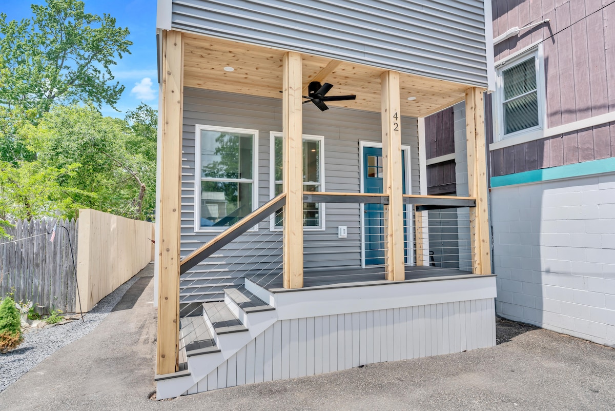 The entrance of a new home features a covered porch with a wooden ceiling and a ceiling fan. Steps lead up to the porch, which is bordered by stylish railing and showcases large windows with white trim.