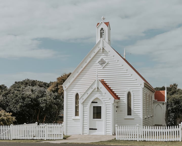  The image shows a charming, small white chapel with a red roof and a picket fence surrounding it. Its traditional architecture features arched windows and a steeple with a bell tower, creating a quaint, peaceful atmosphere. The building is set against a backdrop of greenery and a cloudy sky, adding to its serene, picturesque feel. The overall scene captures the simplicity and beauty of a classic country chapel.