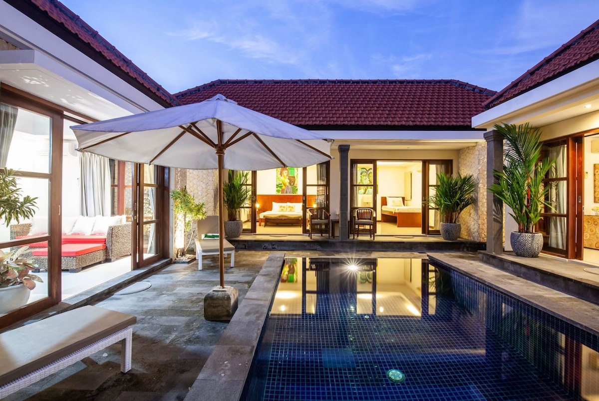A serene pool area is framed by two adjoining villa entrances, showcasing a tranquil blue pool surrounded by stone tiles. An umbrella shades a sunbed, while lush greenery adds a natural touch. Soft lights from the villa hint at a welcoming evening ambiance.