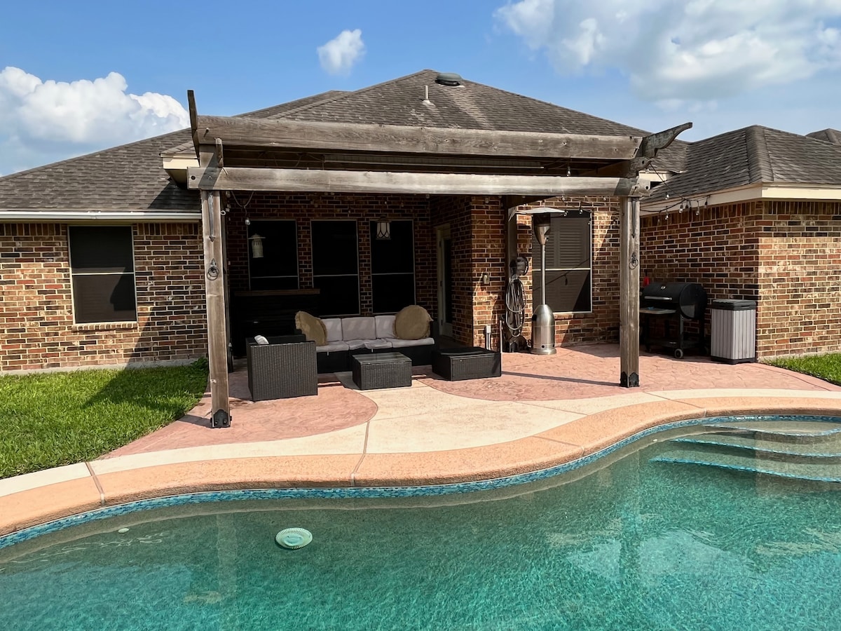 A covered patio area is positioned alongside a swimming pool, featuring a seating arrangement with dark wicker furniture and light cushions. The backdrop displays a brick home with multiple windows, and lush green grass frames the pool's edge, inviting relaxation.