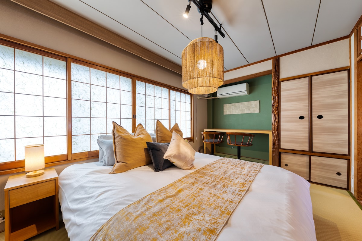 A cozy bedroom is presented, featuring a spacious bed adorned with multiple decorative pillows. A pendant light hangs above, complemented by natural light filtering through traditional paper-paneled windows. A small wooden table is positioned nearby, and storage cabinets are visible, showcasing a blend of textures.