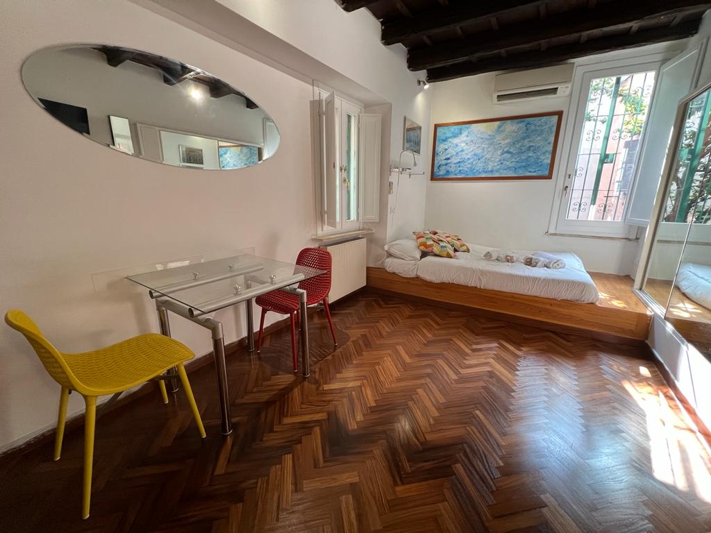 A bright room features a spacious bed with decorative pillows and a glass dining table surrounded by colorful chairs. Natural light enters through large windows, highlighting the herringbone wooden floor and featuring a mirror on the wall that reflects the room's layout.