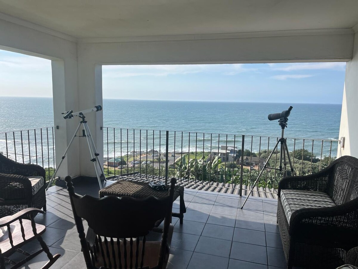 A shaded outdoor seating area features wicker chairs and a swing chair overlooking the ocean. Two telescopes are positioned for viewing, with expansive ocean views visible through the large openings in the walls.