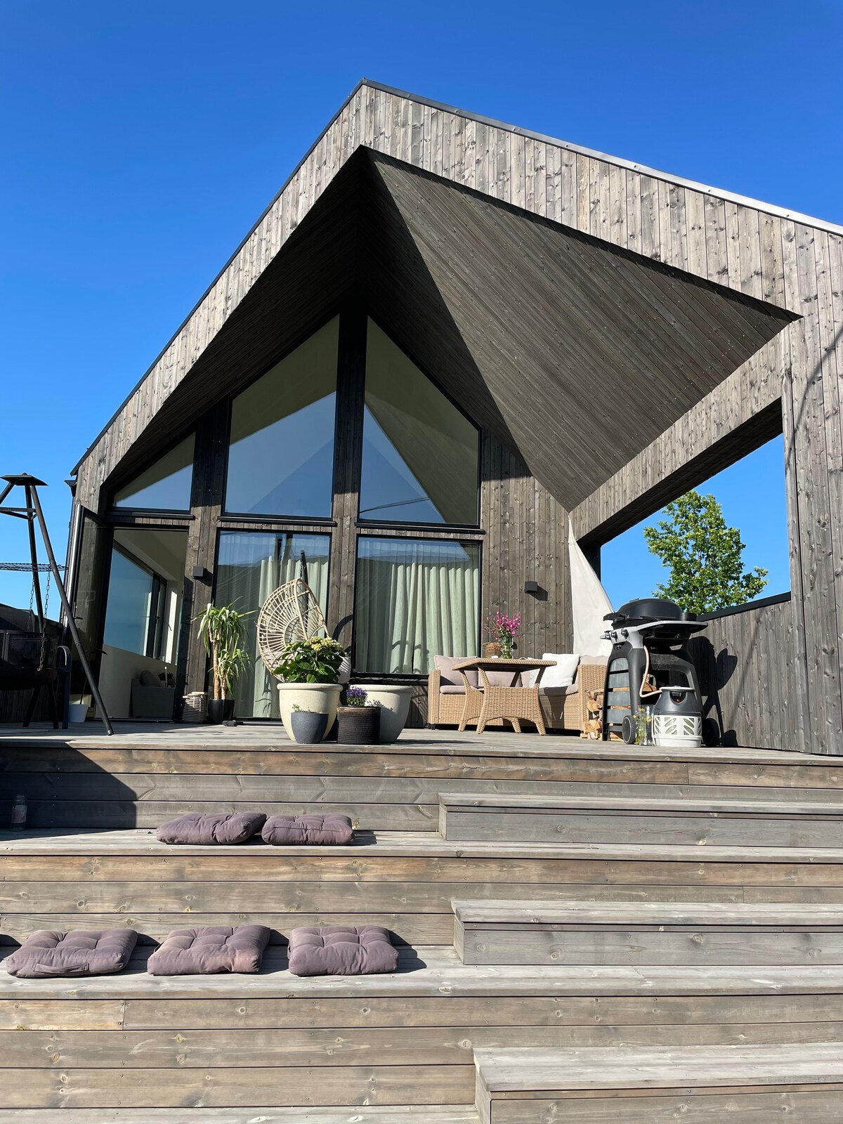 A modern house features an angular wooden exterior and large glass panels, allowing natural light to fill the interior. The entryway includes a set of wooden steps leading up to a spacious deck adorned with potted plants and outdoor furniture, inviting relaxation.