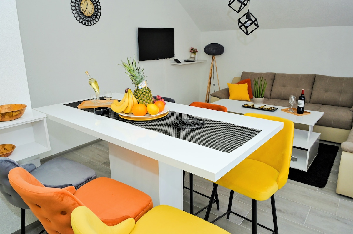 A spacious dining area showcases a bright table with a decorative centerpiece of fresh fruit. Colorful chairs in gray, orange, and yellow surround the table. A comfortable seating area featuring a neutral-colored sofa and a TV in the background enhances the inviting atmosphere.