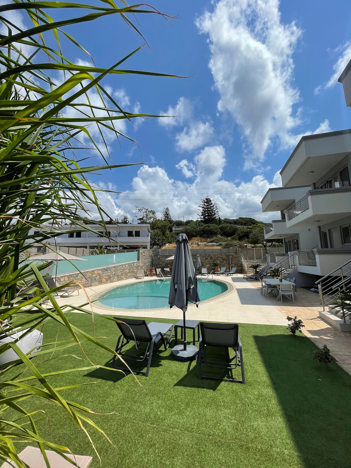 A spacious outdoor area features a shared swimming pool, surrounded by sun loungers and umbrellas. Lush greenery is present, and a manicured lawn adds to the relaxing atmosphere. The property building stands in the background, showcasing a modern architectural design under a bright blue sky.