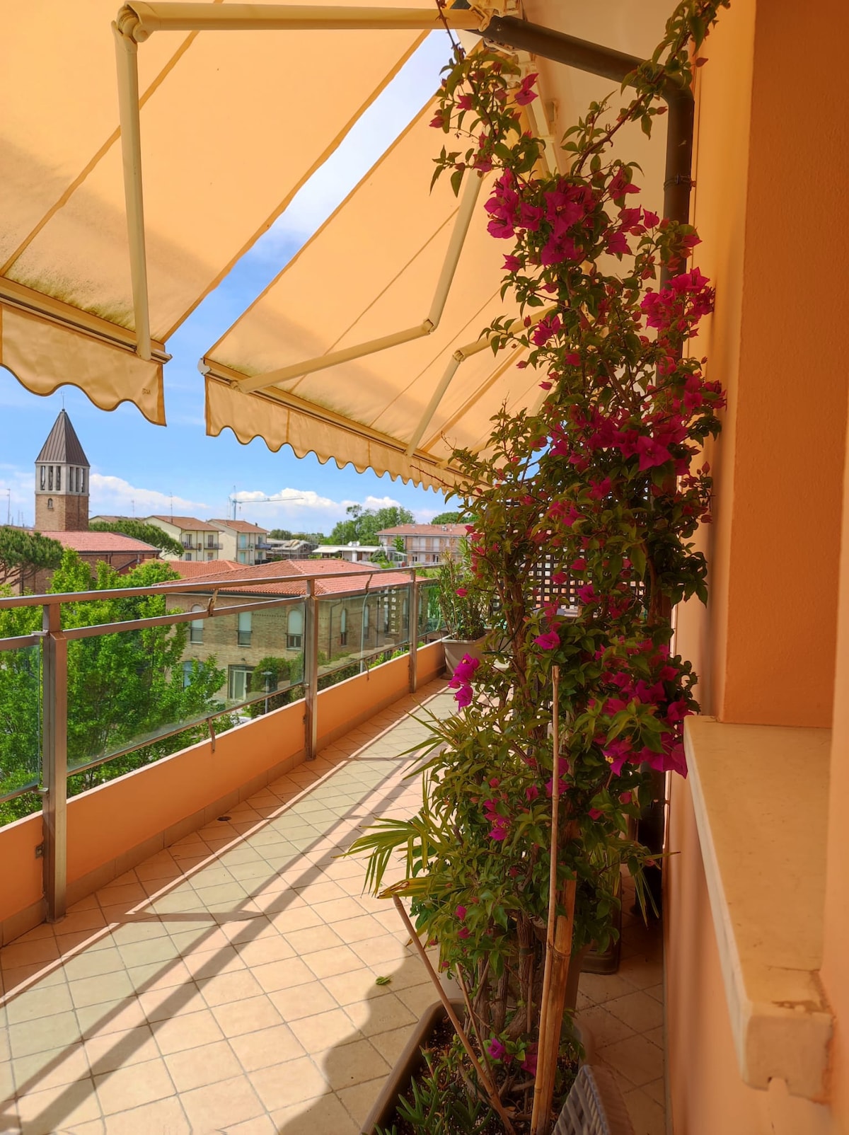 A spacious terrace is highlighted by vibrant bougainvillea and a partially shaded area provided by retractable awnings. The view includes surrounding buildings and greenery, with a bell tower visible in the background. The tiled floor offers a welcoming space for outdoor relaxation or dining.
