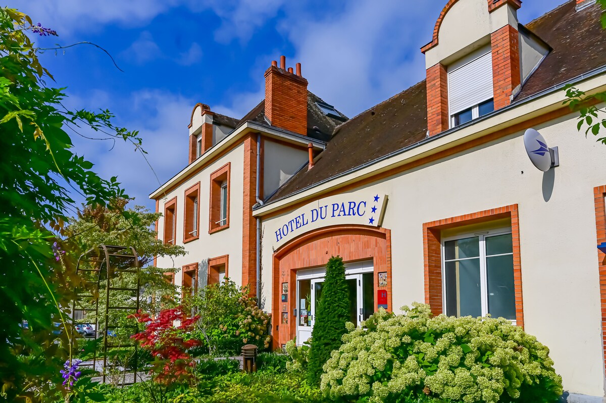 The hotel facade displays a welcoming entrance with the name prominently featured. Flowering plants and shrubs frame the sidewalk, while a mix of brick and stucco materials creates a charming exterior. The blue sky and soft clouds provide a serene backdrop.