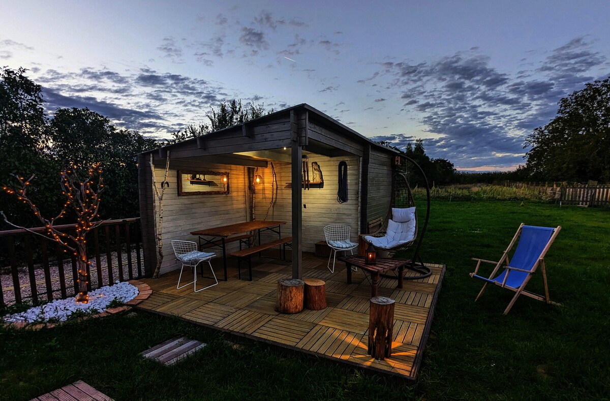 An outdoor wooden terrace is illuminated softly, featuring a large table with seating and a cozy hanging chair. Surrounding greenery and decorative lights enhance the inviting atmosphere, while an open sky offers a backdrop of wispy clouds at dusk.