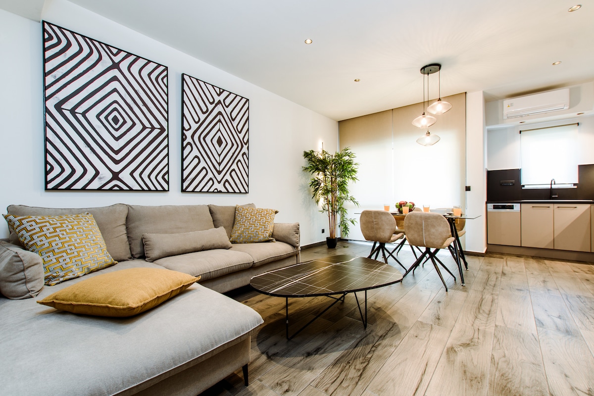 A modern living space features a comfortable sectional sofa adorned with decorative cushions, complemented by a sleek coffee table in front. Two large abstract paintings hang on the wall, while natural light filters through the windows, enhancing the inviting atmosphere. A dining area with a small table is visible, creating a cozy gathering spot.