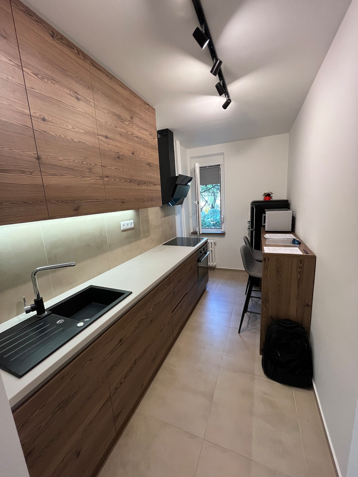 The kitchen displays modern wooden cabinetry with a seamless, clean design. A double sink and sleek black countertop highlight the space. A small dining table is positioned near a window, which offers natural light and views of the outdoors. A chair and a dark backpack are visible.
