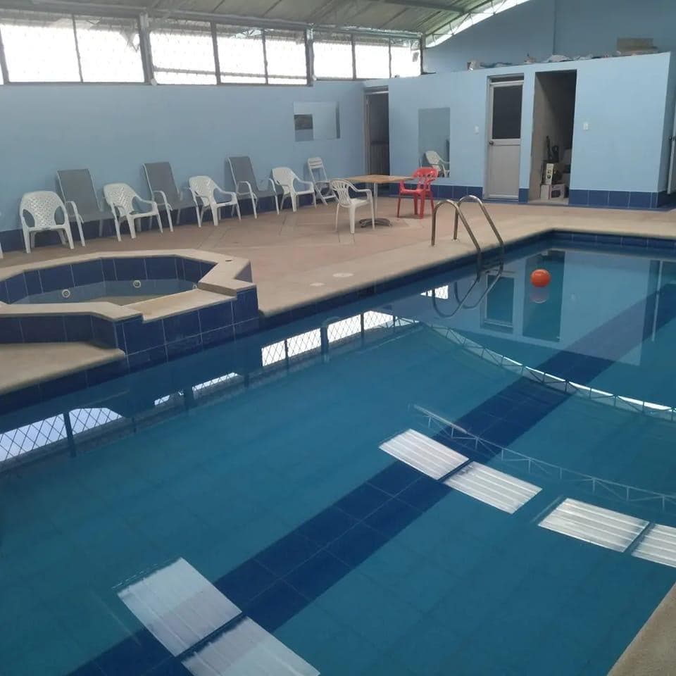 An indoor pool area features clear blue water and a shallow section with a small orange ball floating. Surrounding the pool, multiple white lounge chairs are positioned, with a red table visible in the background. Natural light streams through the overhead windows, adding brightness to the space.