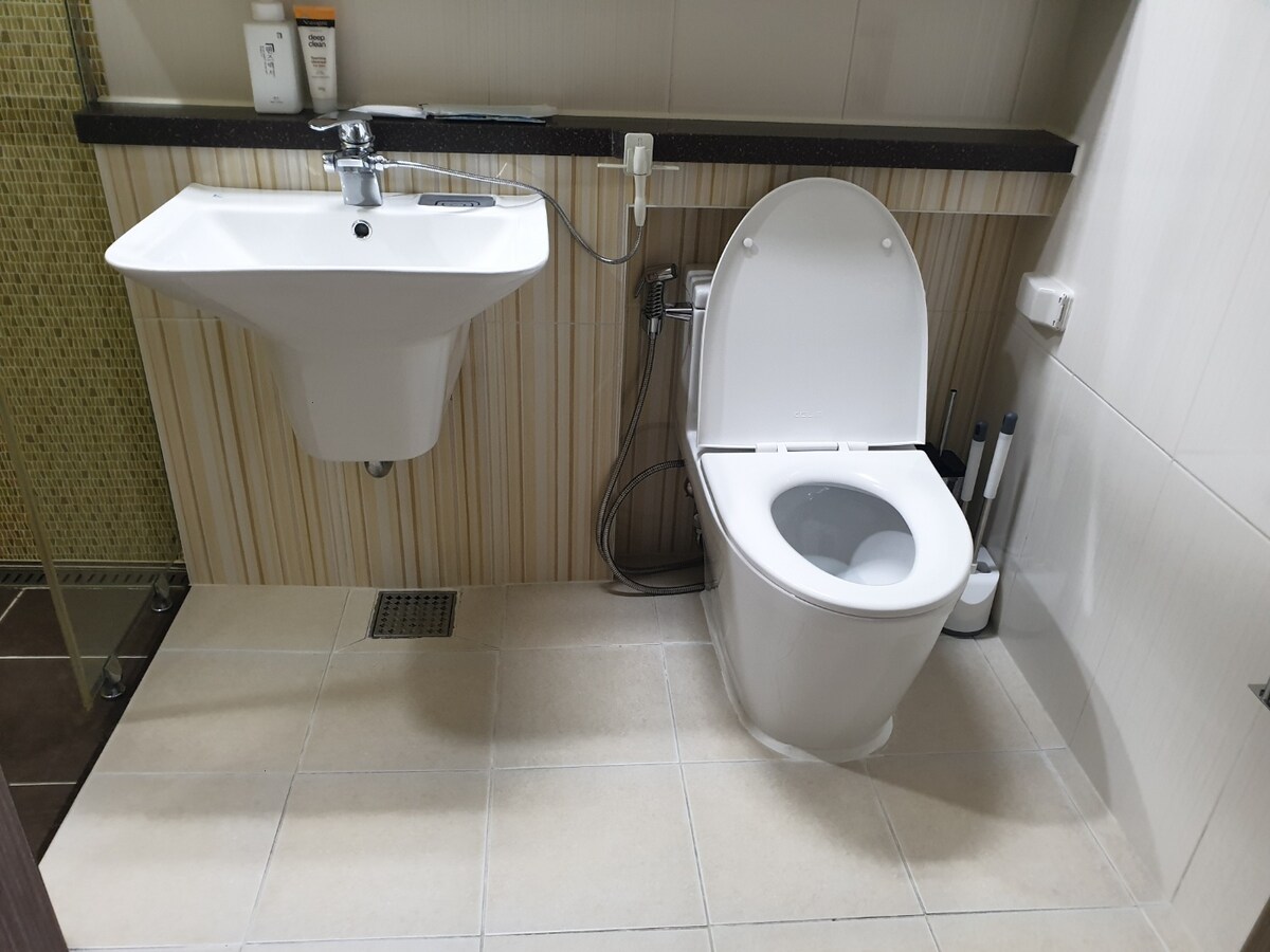 The bathroom features a modern toilet and a wall-mounted sink, both positioned on a tiled floor. Nearby, essential toiletries are visible on a shelf, and a shower area can be seen in the background, enclosed with glass.