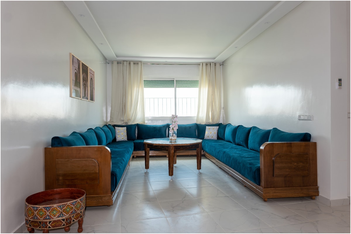 A spacious living area is showcased with a large teal sectional sofa, offering ample seating. A wooden coffee table centers the space, complemented by decorative throw pillows. Natural light filters through the windows, which are framed by soft white curtains.