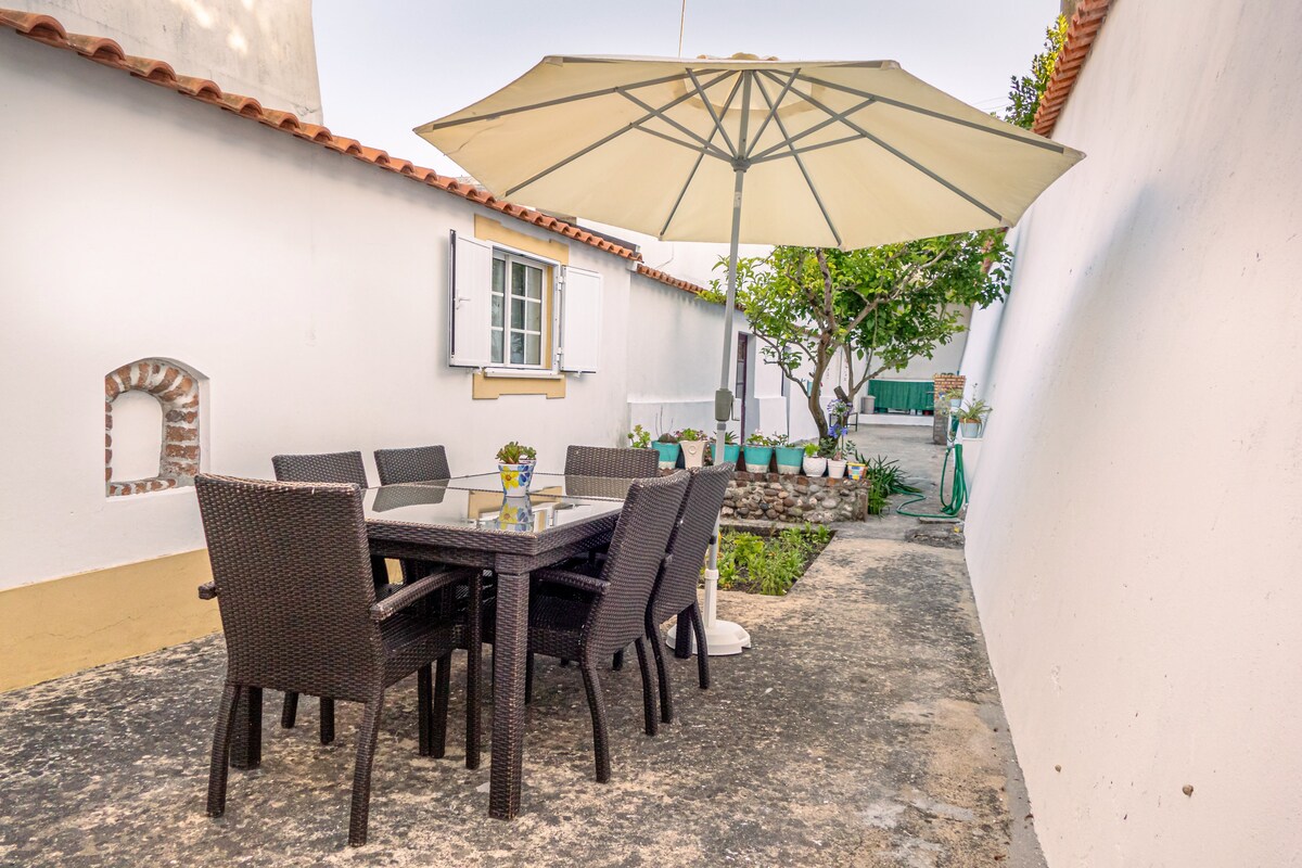 An outdoor patio area is displayed, featuring a dark wicker dining table surrounded by six matching chairs. A large umbrella provides shade, while greenery is visible in the background. A small flower pot adds a decorative touch to the table, enhancing the inviting space.
