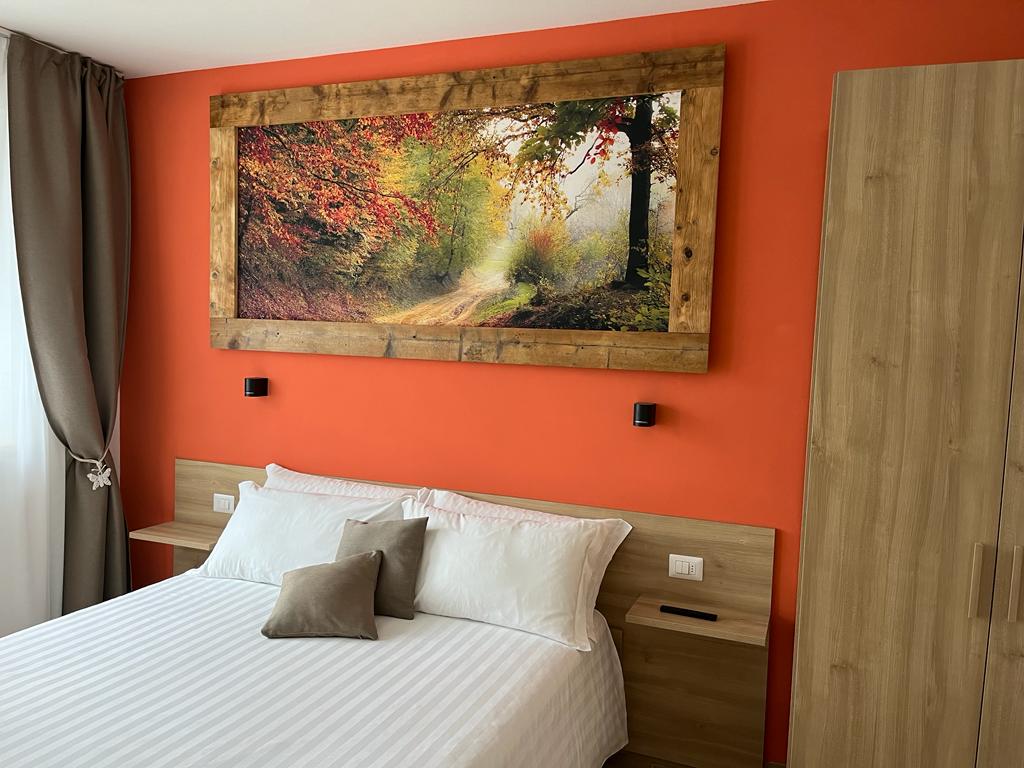 A serene sleeping area features a spacious bed with white linens and soft pillows. An orange accent wall is complemented by a large framed photograph of a forest path. Modern bedside tables and a wooden wardrobe are visible, enhancing the cozy atmosphere.