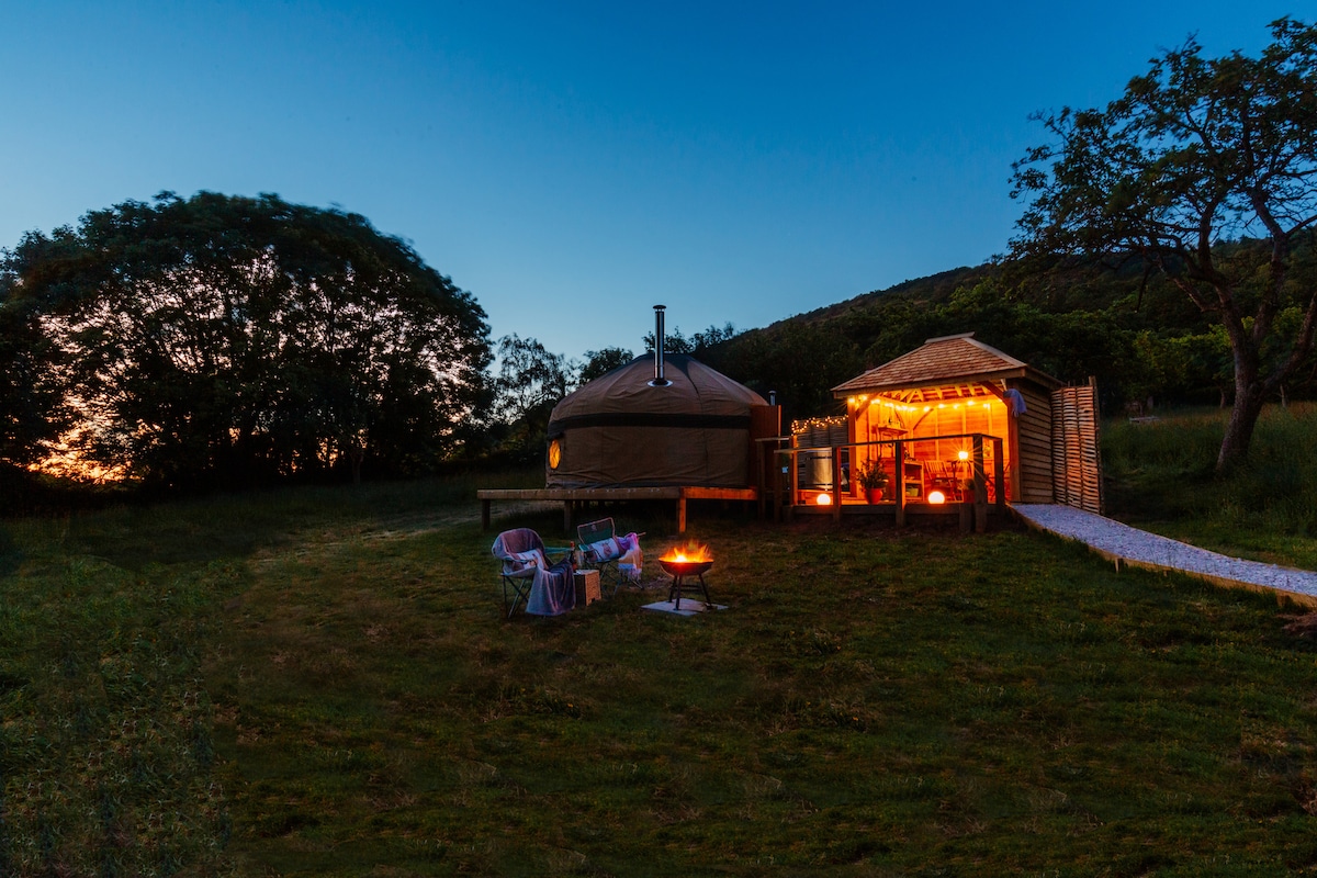 A spacious yurt is nestled in a green landscape, with a warm glow illuminating the outdoor gazebo. Comfortable seating is arranged around a firepit, creating a welcoming atmosphere for evening gatherings. The scene is set against a twilight sky and surrounding trees.