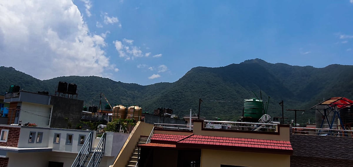 A panoramic view of the surrounding hills is captured, showcasing lush greenery under a bright blue sky with scattered clouds. Rooftops of nearby buildings are visible in the foreground, adding a sense of scale to the mountainous landscape.
