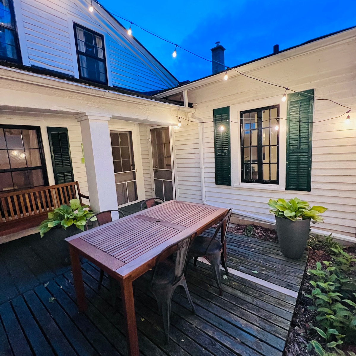 An outdoor dining area features a wooden table surrounded by chairs, illuminated by string lights overhead. Green plants are placed in the corners, adding a natural touch to the wood-planked patio, while the white house exterior is complemented by dark green shutters.