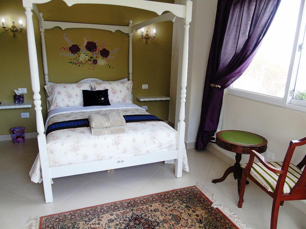 A 4-poster bed is positioned centrally in the room, adorned with floral bedding. A delicate rose mural is painted above the bed, while two sconces provide soft lighting. A small round table sits beside a striped chair, and a large window offers natural light and views of the outside.