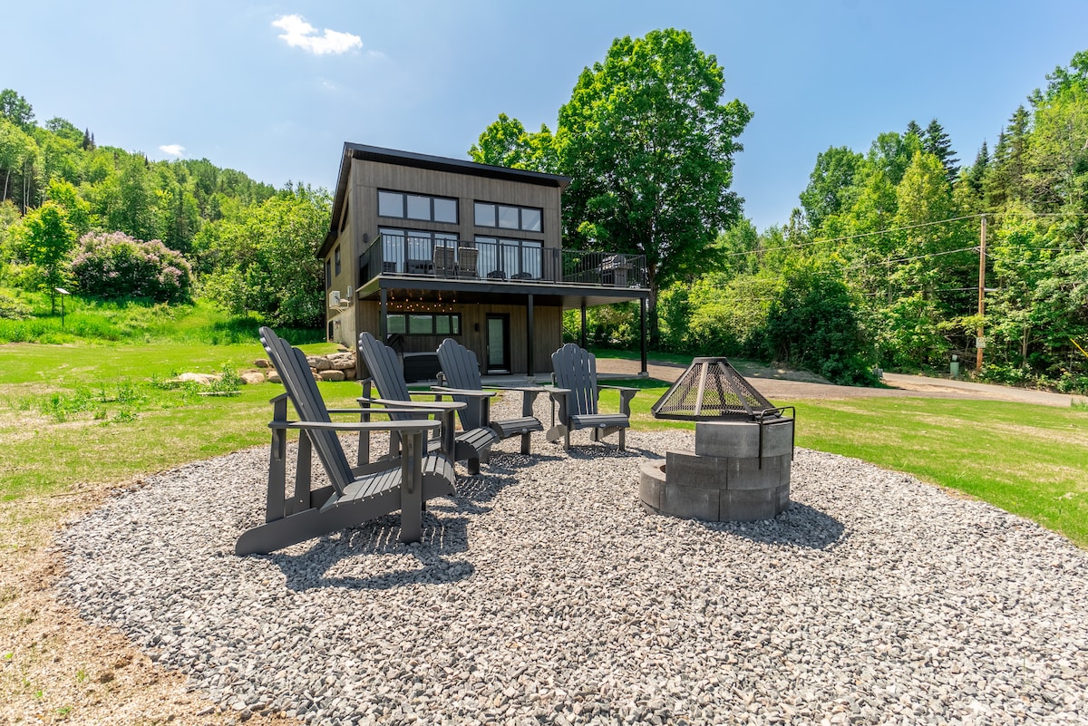 A spacious outdoor area features a circular arrangement of Adirondack chairs surrounding a stone fire pit. The chalet, set against a backdrop of greenery, is visible in the distance, complemented by a clear blue sky.