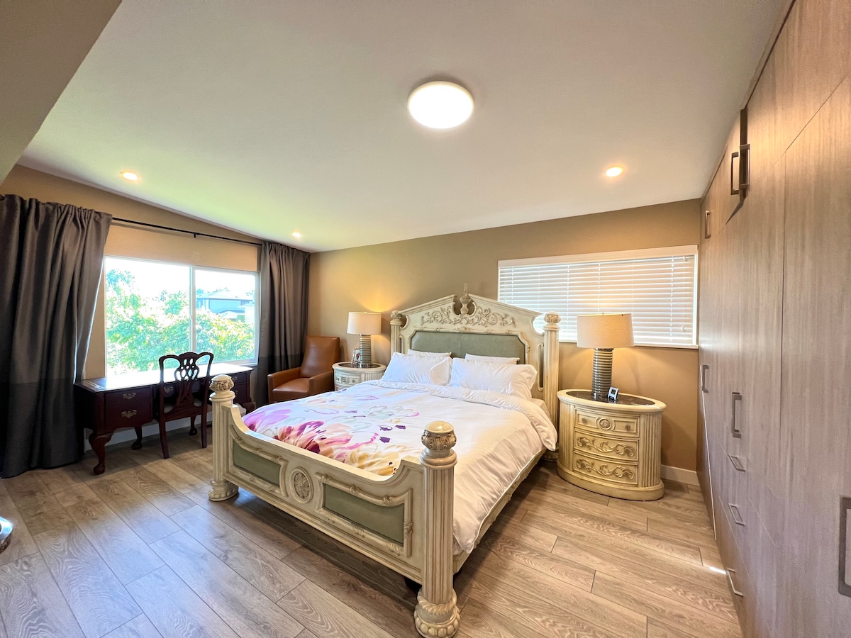 A spacious suite features an oversized bed with decorative elements against a soft backdrop. Natural light filters through large windows with dark curtains, illuminating a cozy sitting area with a chair and a vintage-style desk. Ample wardrobe space is integrated along the wall.