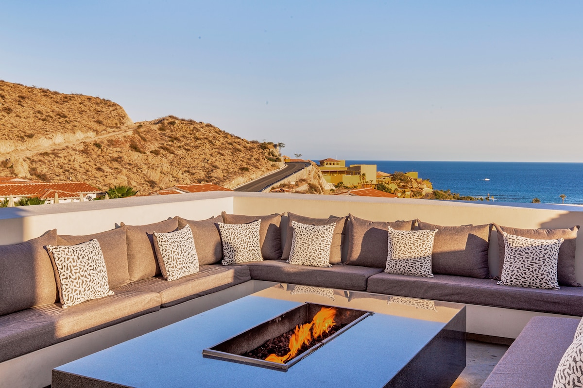 A spacious outdoor seating area features a large sectional sofa with decorative cushions, surrounding a fire pit. Panoramic views of the sea and coastline are visible, providing a serene atmosphere for relaxation and enjoyment.