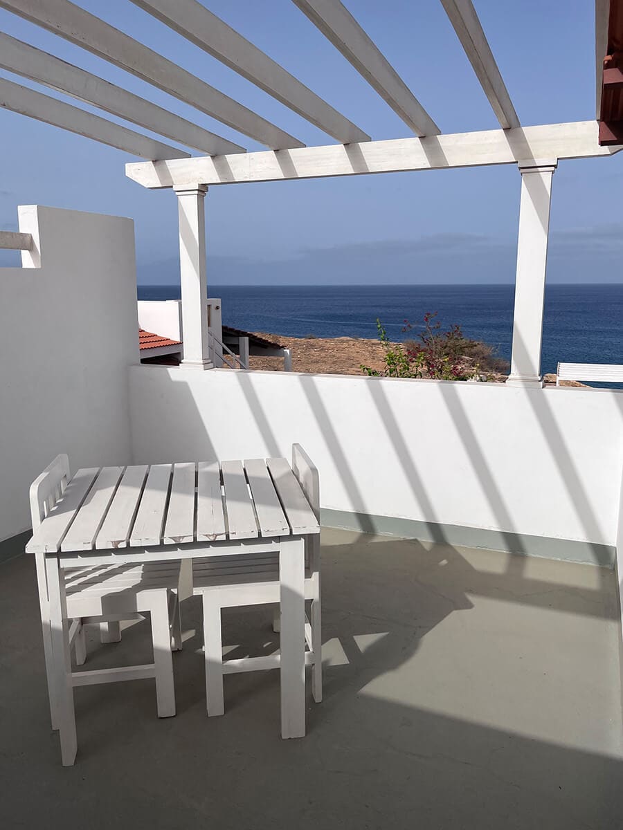 A small terrace features a white wooden table and two matching chairs, positioned under a slatted pergola. The ocean can be seen in the background, with a clear blue sky providing a serene backdrop.
