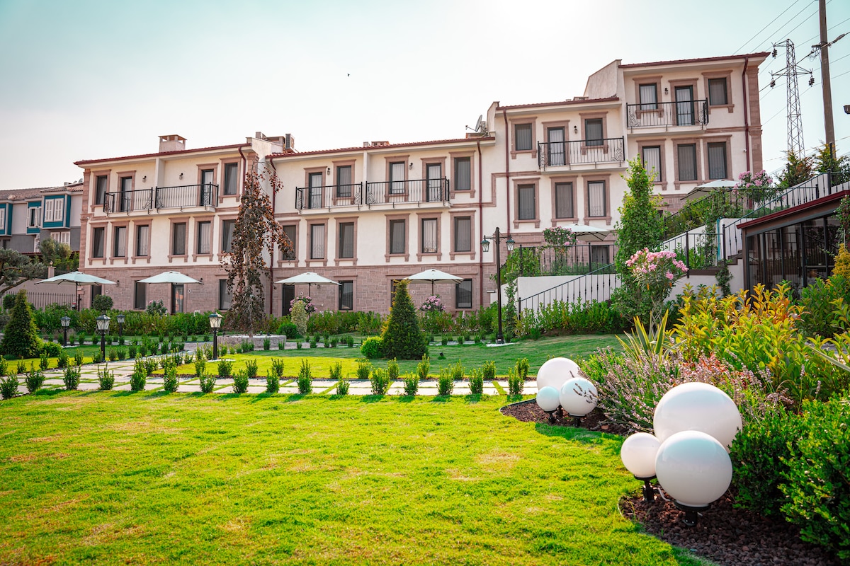 A contemporary hotel building is set against a clear sky, featuring multiple balconies that overlook a manicured garden. Lush greenery surrounds the property, with landscaped paths and decorative stone accents enhancing the tranquil atmosphere.