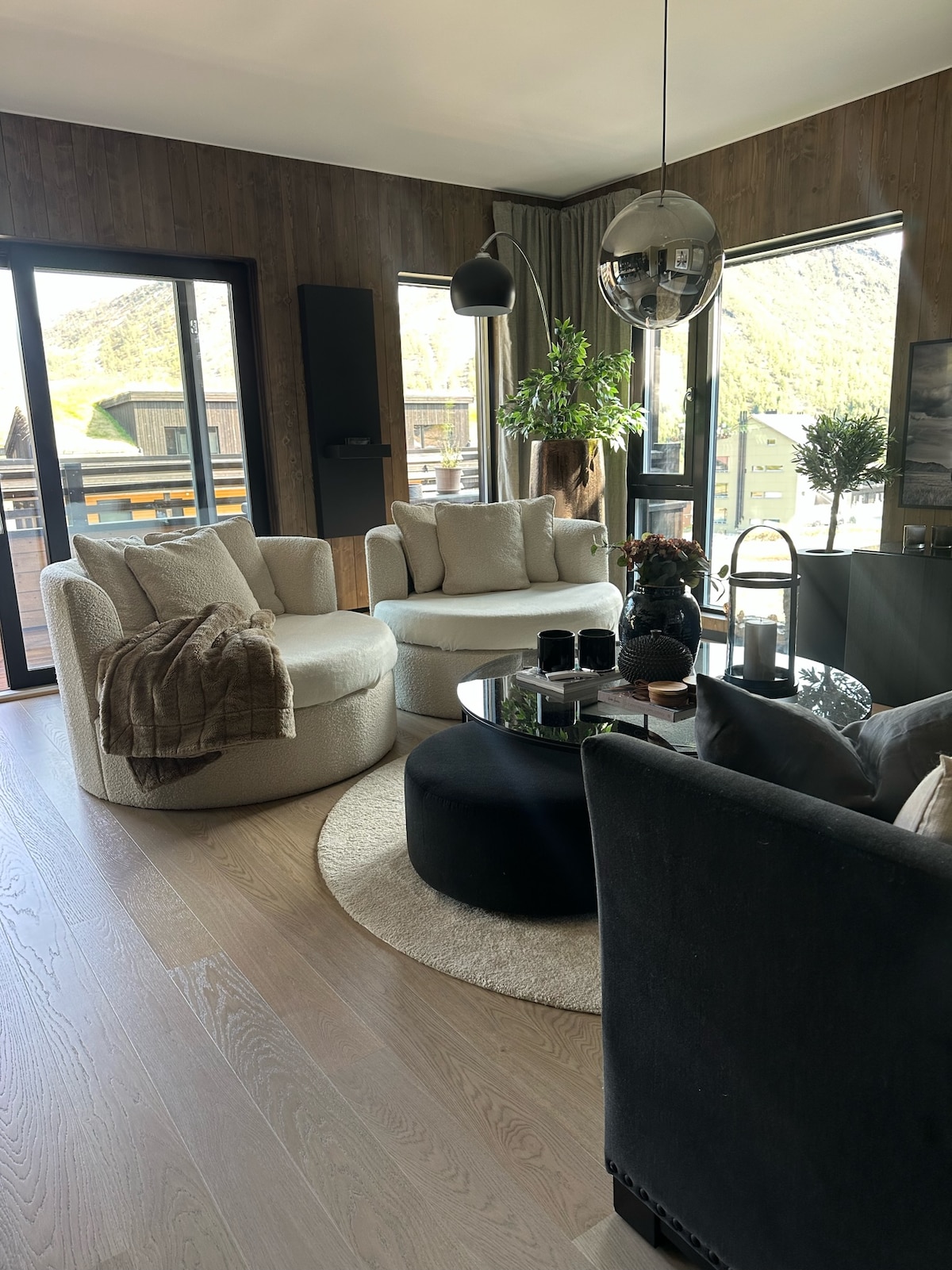 A modern living area features soft, circular chairs in light fabrics alongside a round black coffee table. Large windows provide views of the outdoors, with a potted plant bringing greenery into the space. Natural light enhances the inviting interior designed for comfort.