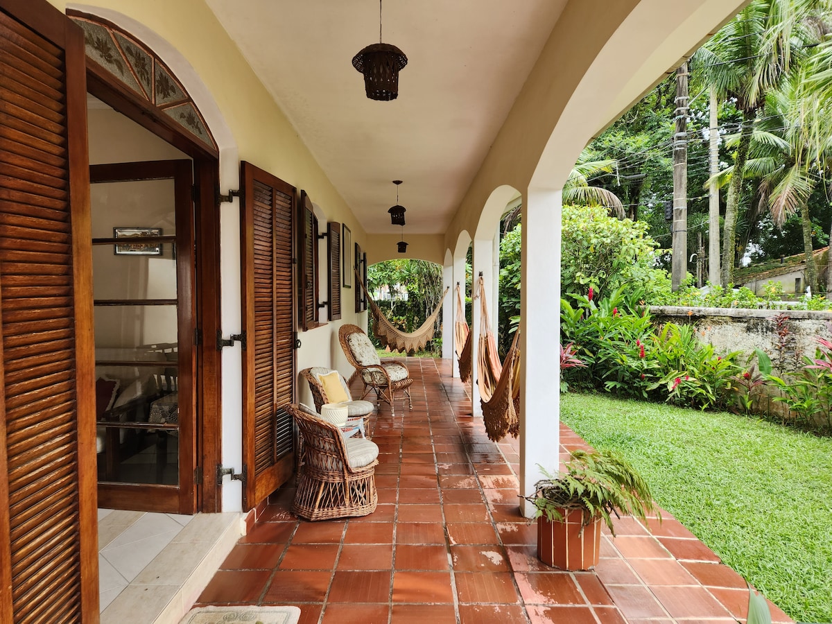 A covered porch is displayed with terracotta tiles and features woven chairs positioned near decorative plants. Hammocks are suspended between the columns, inviting relaxation. Lush greenery and palm trees are visible beyond the porch, creating a serene outdoor ambiance.