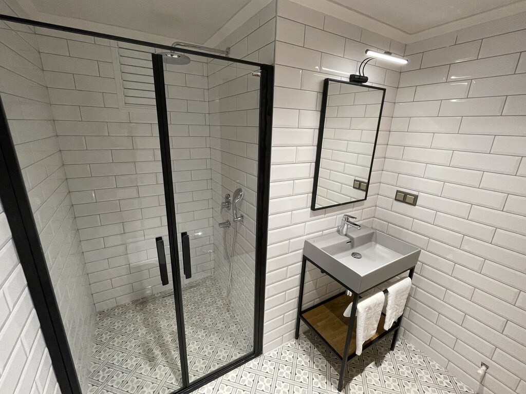 A modern bathroom is presented with sleek glass shower doors and a minimalist sink area. White subway tiles create a clean backdrop, complemented by patterned floor tiles. A large mirror and wall-mounted light add functionality to the space, with neatly arranged towels on a rack.