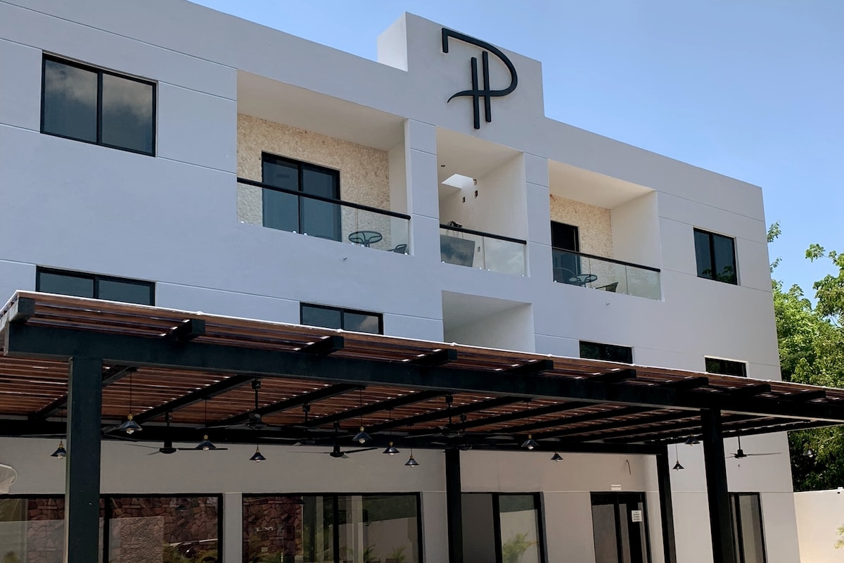 A modern building exterior is presented, showcasing clean lines and a minimalist design. Large windows allow natural light to fill the interior. A shaded outdoor patio with wooden beams provides seating areas, and the building features a prominent logo above the entrance.