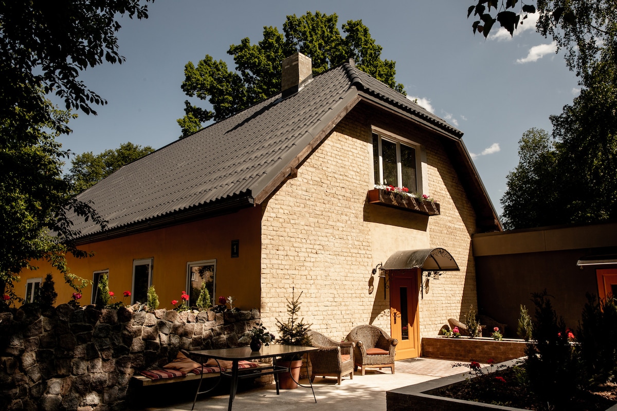 The exterior of a charming building is displayed, featuring a stone facade and a sloped roof. The entrance is highlighted by a covered awning and surrounded by potted flowers. A stone patio offers spaces for relaxation with lounge furniture and natural greenery in the background.
