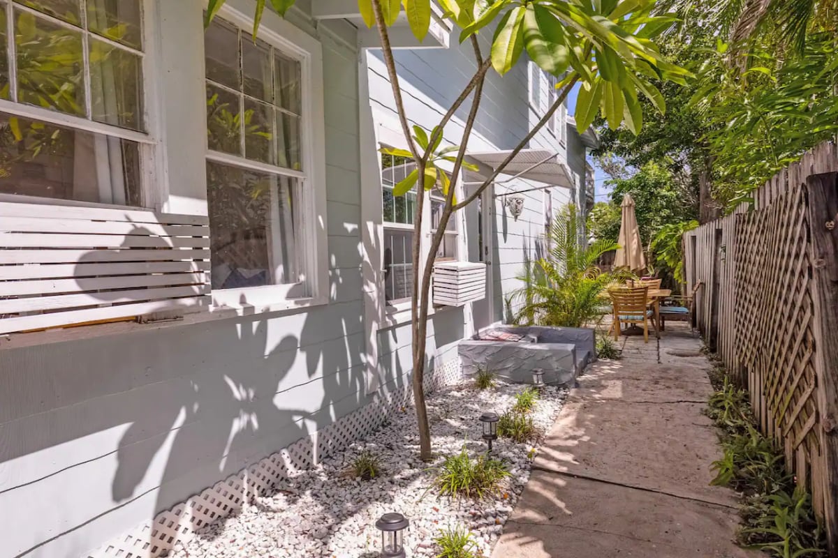 A tranquil pathway is lined with white gravel and lush greenery, leading past a shade tree towards a seating area. Sunlight filters through the leaves, casting gentle shadows on the siding of the building and illuminating an outdoor dining set.