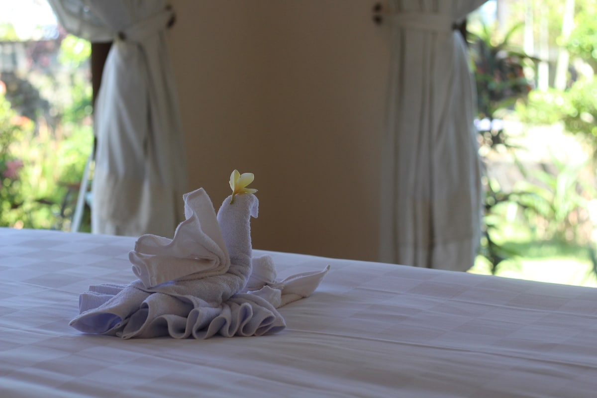 A neatly arranged towel sculpture is placed on a comfortable bed, adorned with a fresh flower. Soft natural light filters through the large open curtains, highlighting the vibrant greenery visible outside the windows.