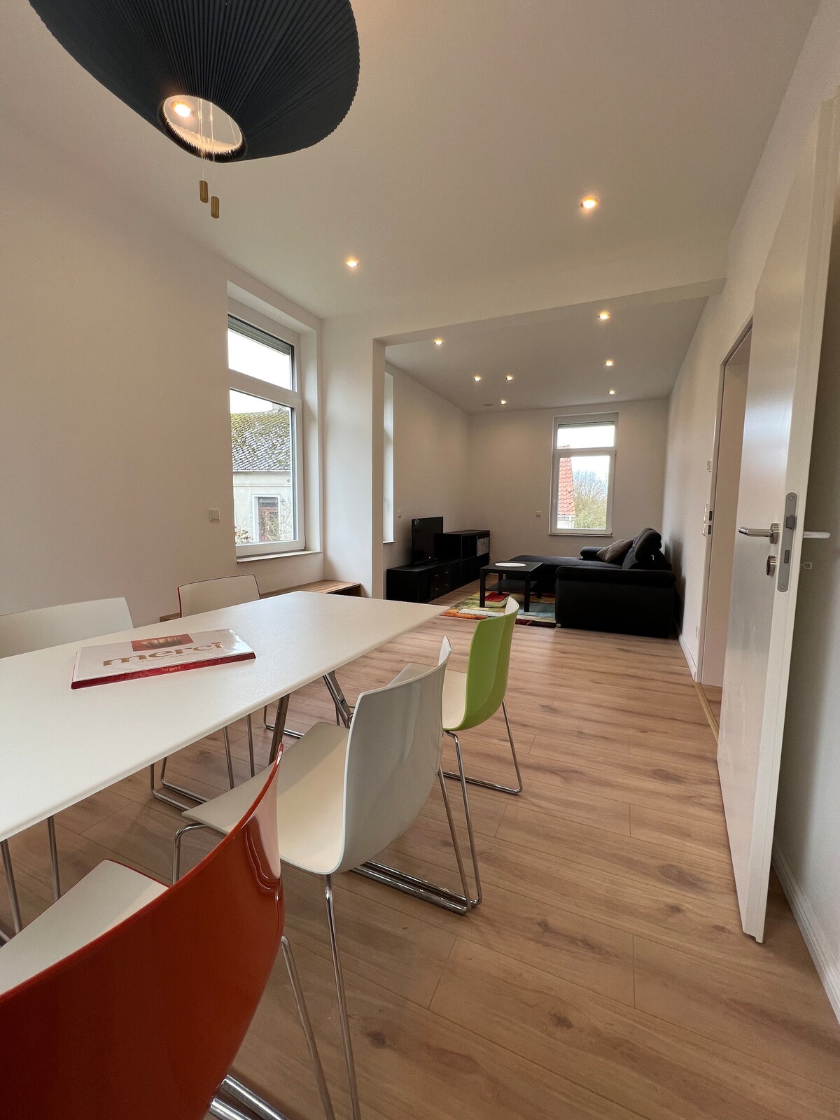 A dining area is showcased with a long table surrounded by colorful chairs in various shades. Large windows allow natural light to brighten the space. A cozy seating arrangement with a dark sofa is visible in the background, contributing to a welcoming environment.