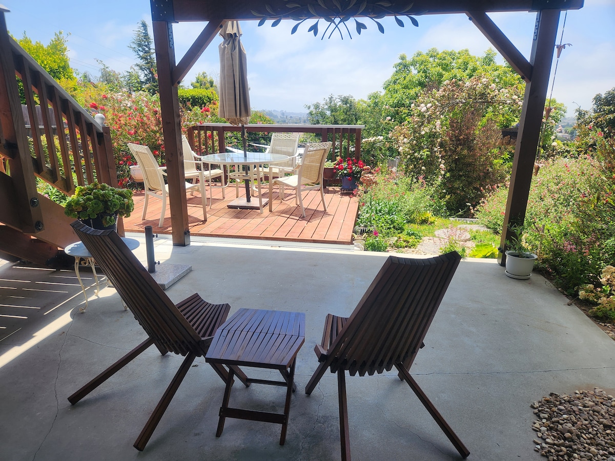 A shaded outdoor patio area features two wooden lounge chairs positioned toward a small table. A staircase leads to a wooden deck adorned with a dining table and chairs, surrounded by vibrant greenery and blossoming flowers, creating a serene outdoor environment.