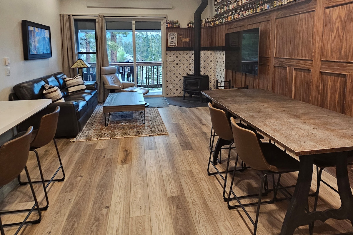 The open living area showcases a comfortable leather sofa and a wooden coffee table. A dining table with multiple chairs is positioned nearby. A wood-burning stove is located against the wall, with a large window offering views of the outside trees and light filtering into the room.