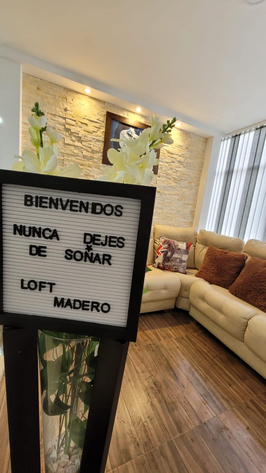 A welcome sign introduces the space, surrounded by elegant flowers. A cozy seating area features a light-toned sectional sofa, adorned with decorative pillows. Soft lighting enhances the stone accent wall, while large windows allow natural light to fill the room.