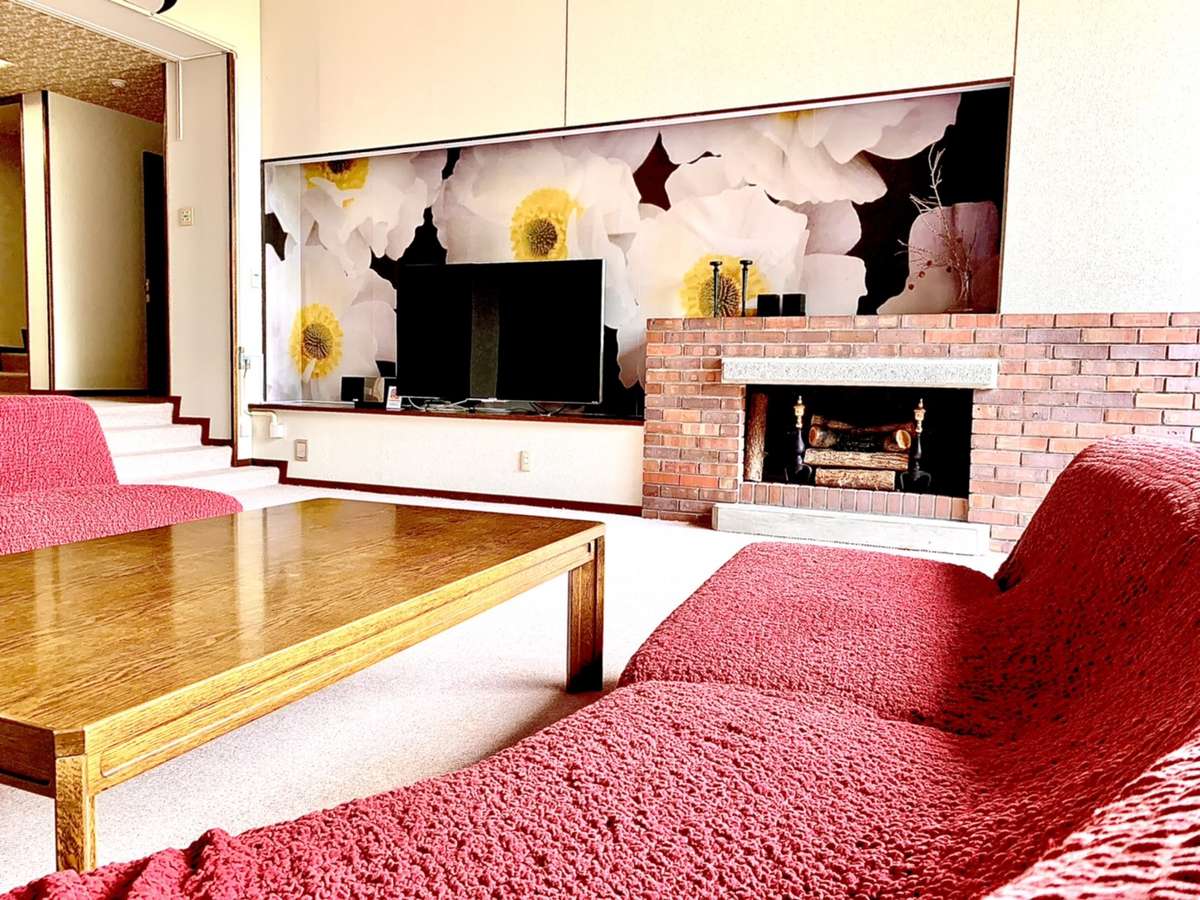 A spacious living room features a flat-screen television mounted on a wall adorned with floral wallpaper. A wooden coffee table sits in front of a cozy seating area with deep red couches, while a brick fireplace adds warmth to the space.