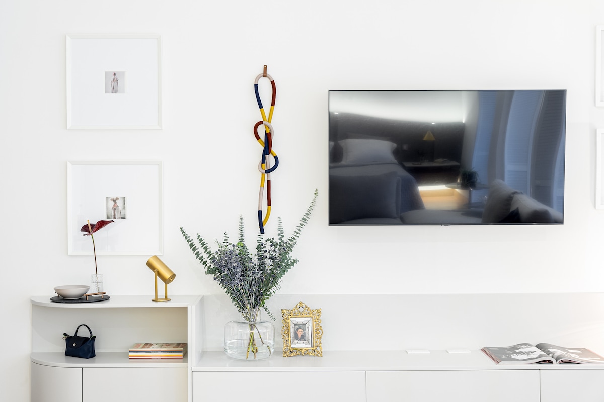 A modern wall displays a television mounted above a sleek console. Framed artwork in light-colored frames adorns the wall. A decorative piece featuring colorful ribbons hangs alongside. A small lamp and a vase with greenery are placed on the console, adding subtle detail to the setting.