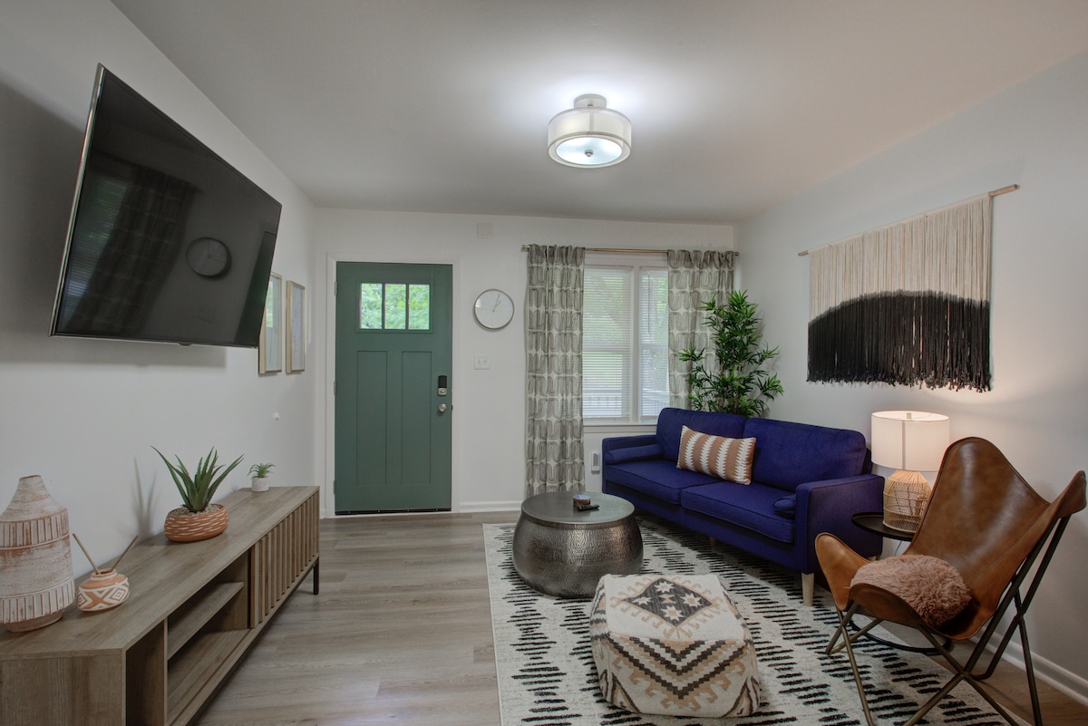 A modern living room features a deep blue sofa and a circular coffee table. A large flat-screen TV is mounted on the wall, and a charming lamp casts a warm light. Natural light filters through curtains, and decorative elements include a plant and textured wall hanging.