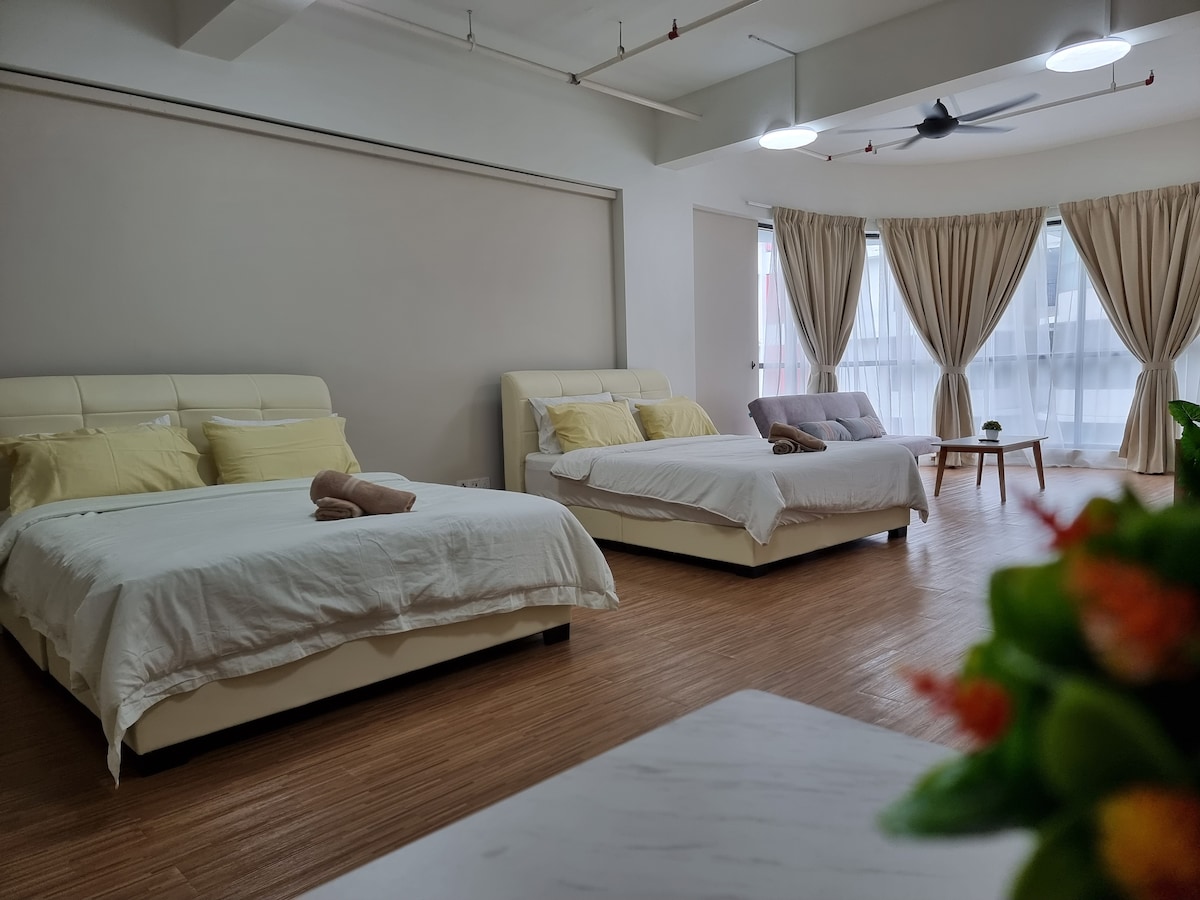 The room features two spacious beds with light-colored linens. Natural light filters through the large windows, which are dressed with soft curtains. A decorative plant is visible in the foreground, adding a touch of greenery to the relaxing space.