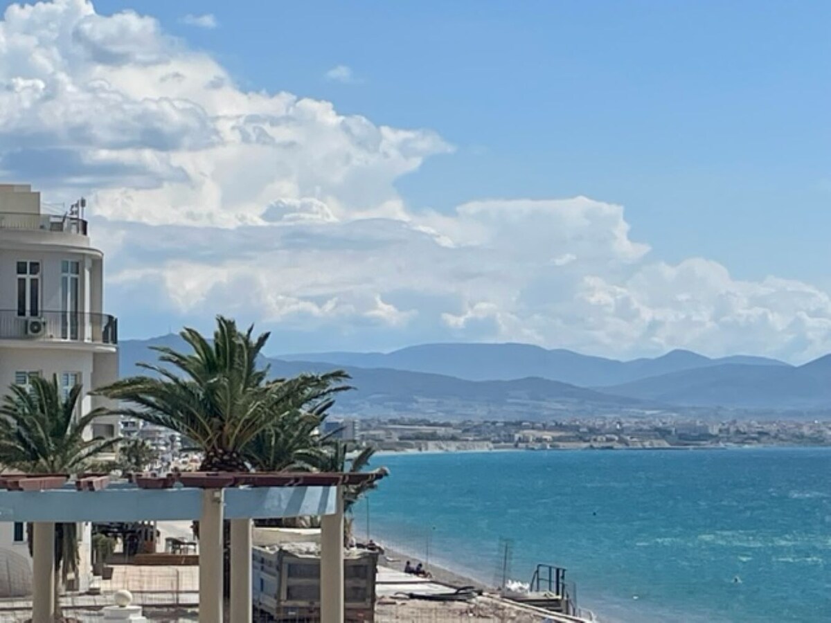 The image captures a coastal view featuring vibrant palm trees and clear turquoise waters. The distant mountains provide a scenic backdrop under a partly cloudy sky, creating a serene atmosphere. The beachfront promenade and neighboring buildings are visible, enhancing the coastal charm.