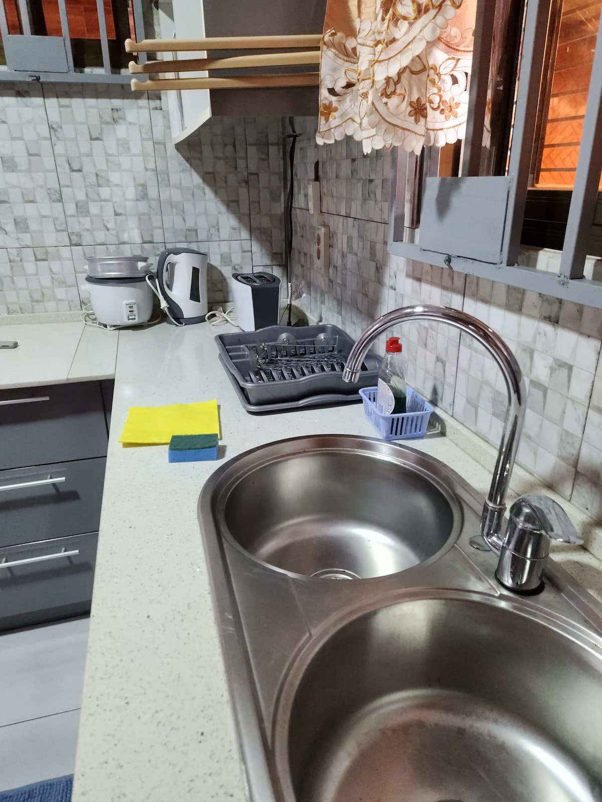 The kitchen features a double sink, complemented by a modern countertop and cabinetry. A drying rack is positioned beside a kettle and a rice cooker. Cleaning supplies are neatly arranged, and sunlight enters through a curtain-adorned window, enhancing the workspace.